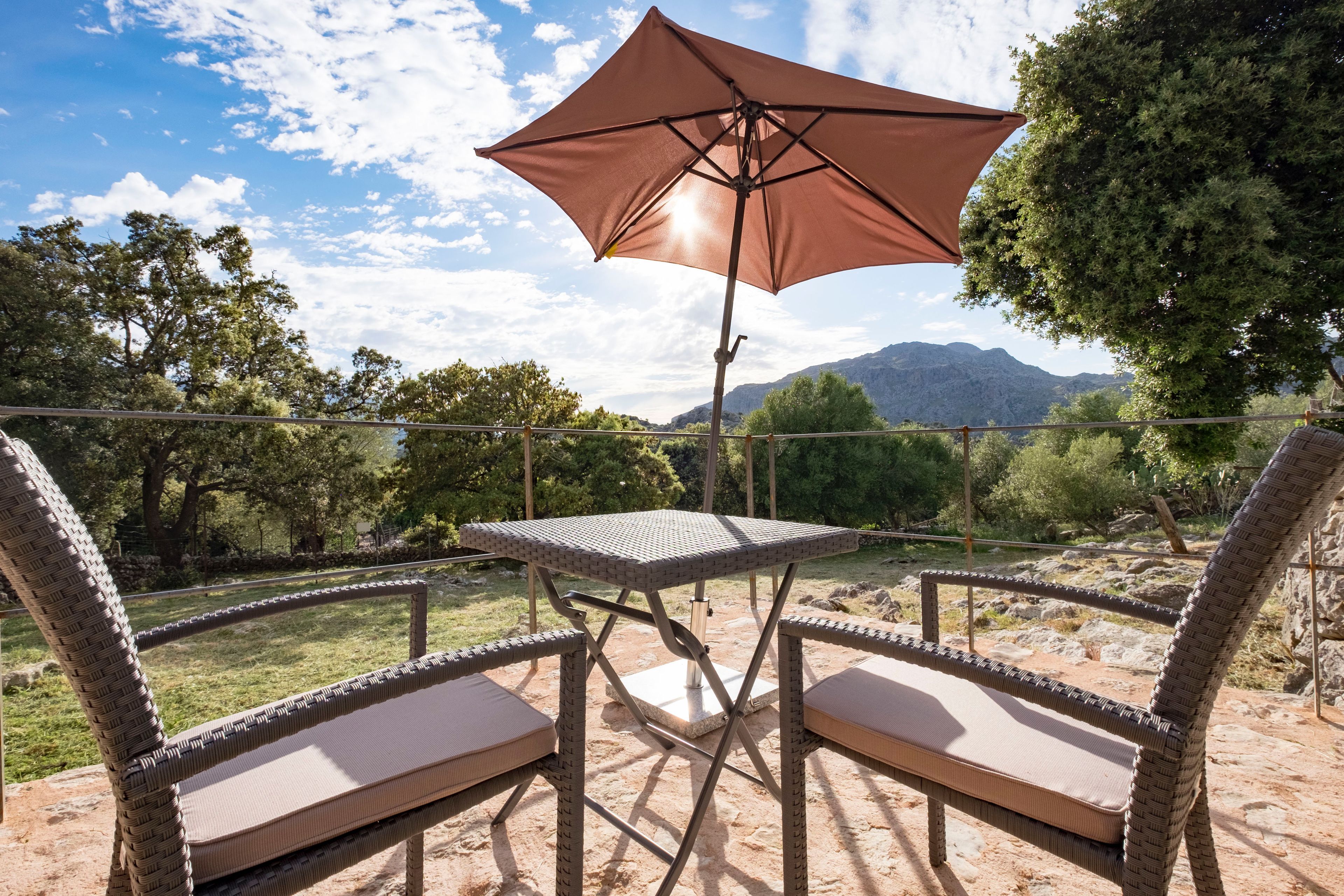 Een terras buiten met rieten stoelen en een tafel onder een grote parasol, met uitzicht op een bergachtig landschap.