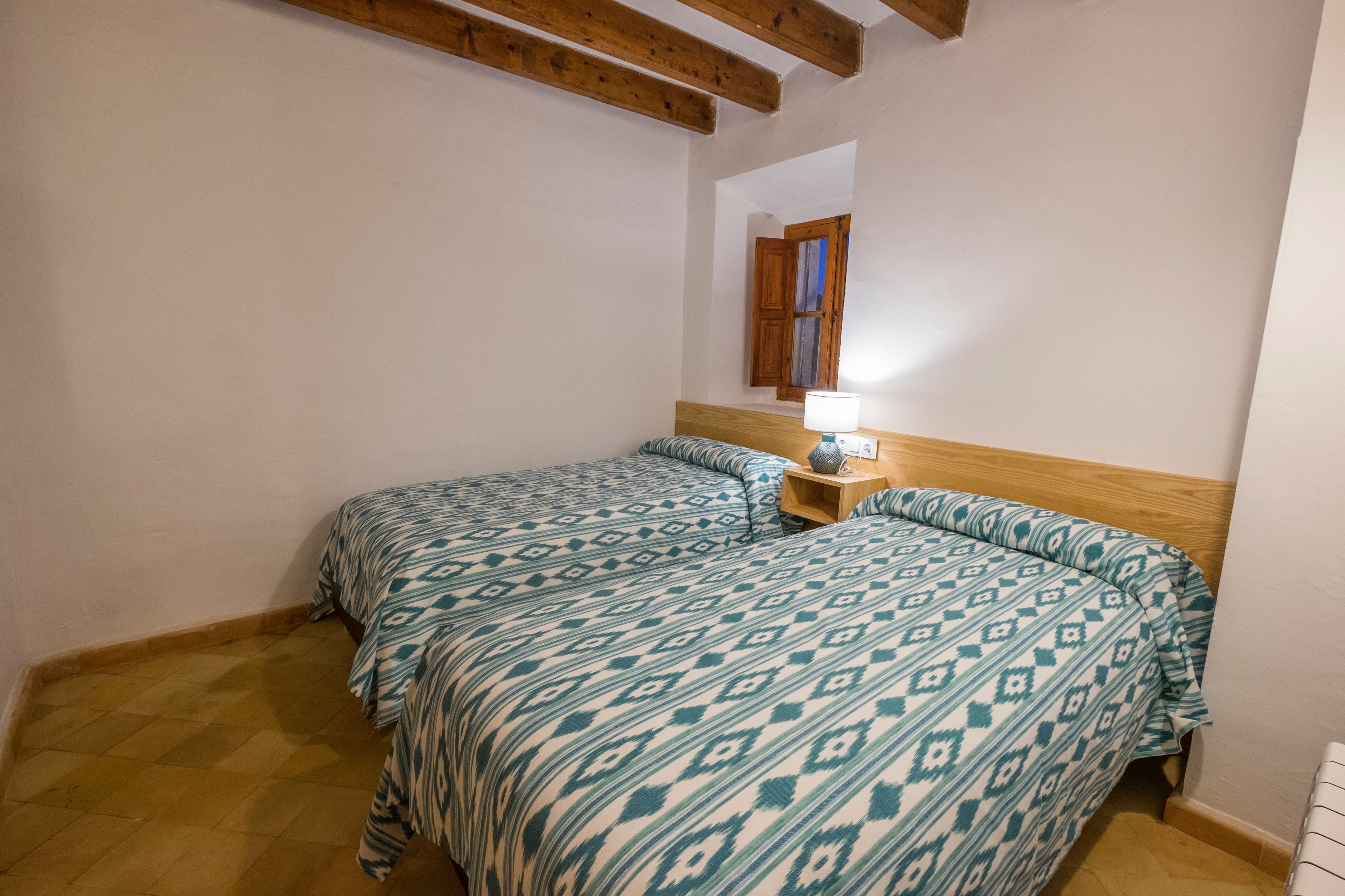 A bedroom with two beds, wooden beams on the ceiling, a nightstand with a lamp, and a window with wooden shutters.