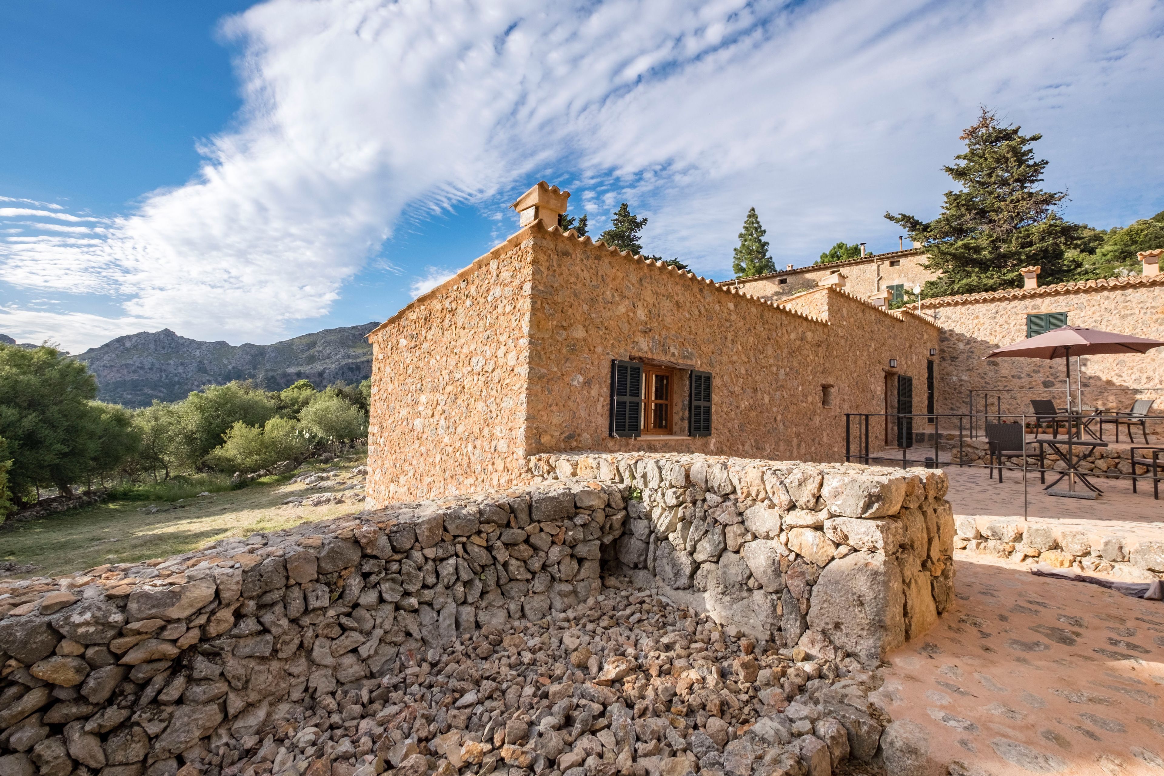 A stone building with a tiled roof, outdoor seating area, and a stone wall in the foreground.