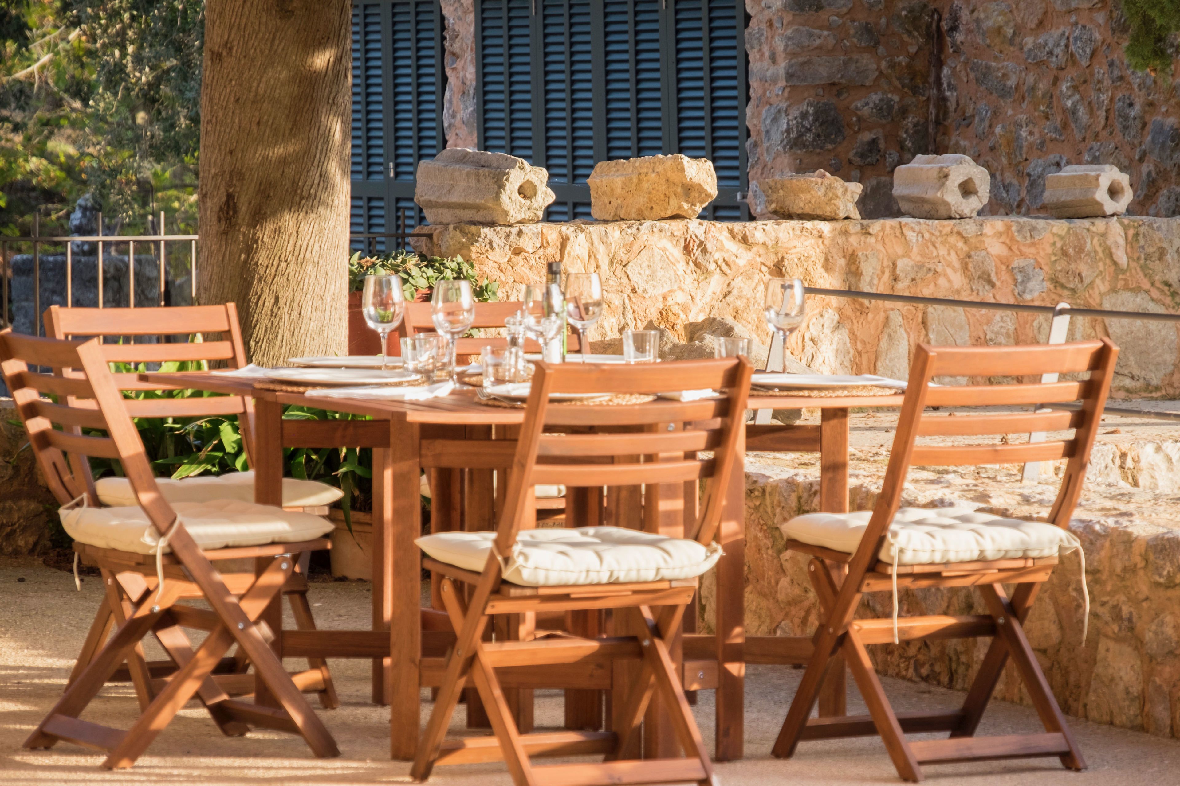 An outdoor dining area with wooden tables and chairs, stone walls, and a tree.