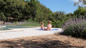 Two children sitting by a poolside with a natural landscape in the background.