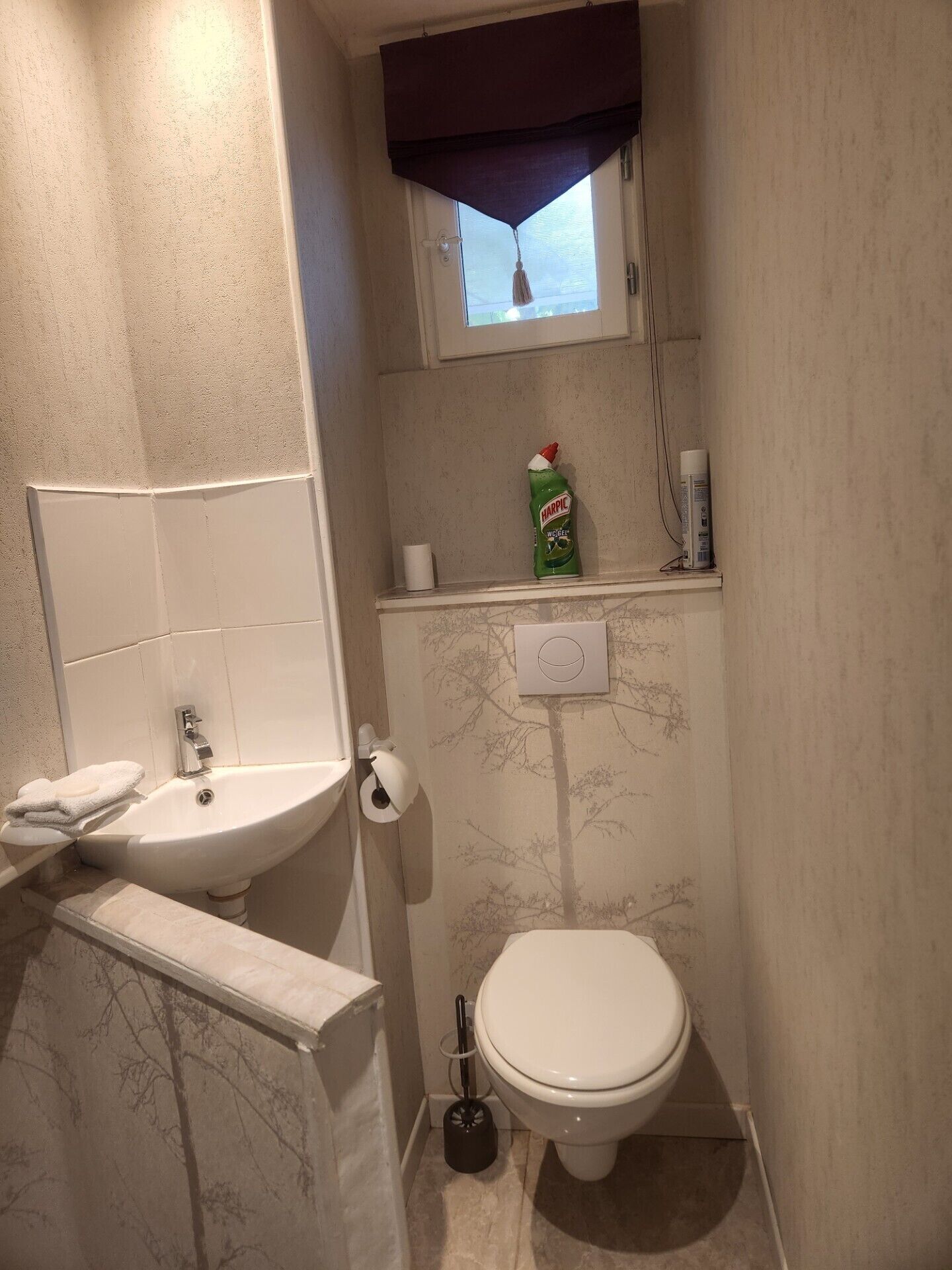 Un baño pequeño con lavamanos, inodoro y ventana con un visillo oscuro.