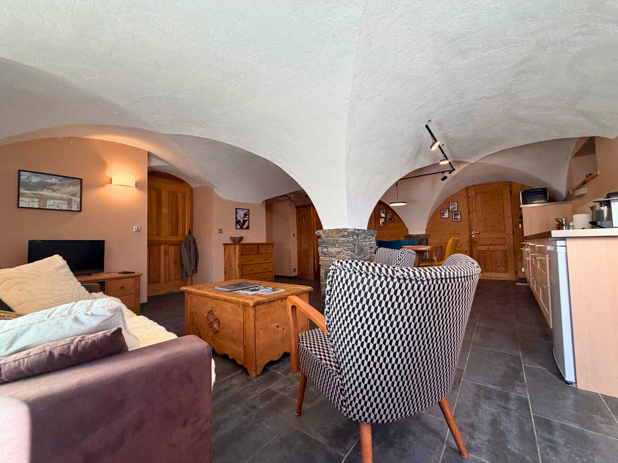 A cozy living room with a sofa, a patterned armchair, a wooden coffee table, and a television. There is a kitchen area with a stove and cabinets.