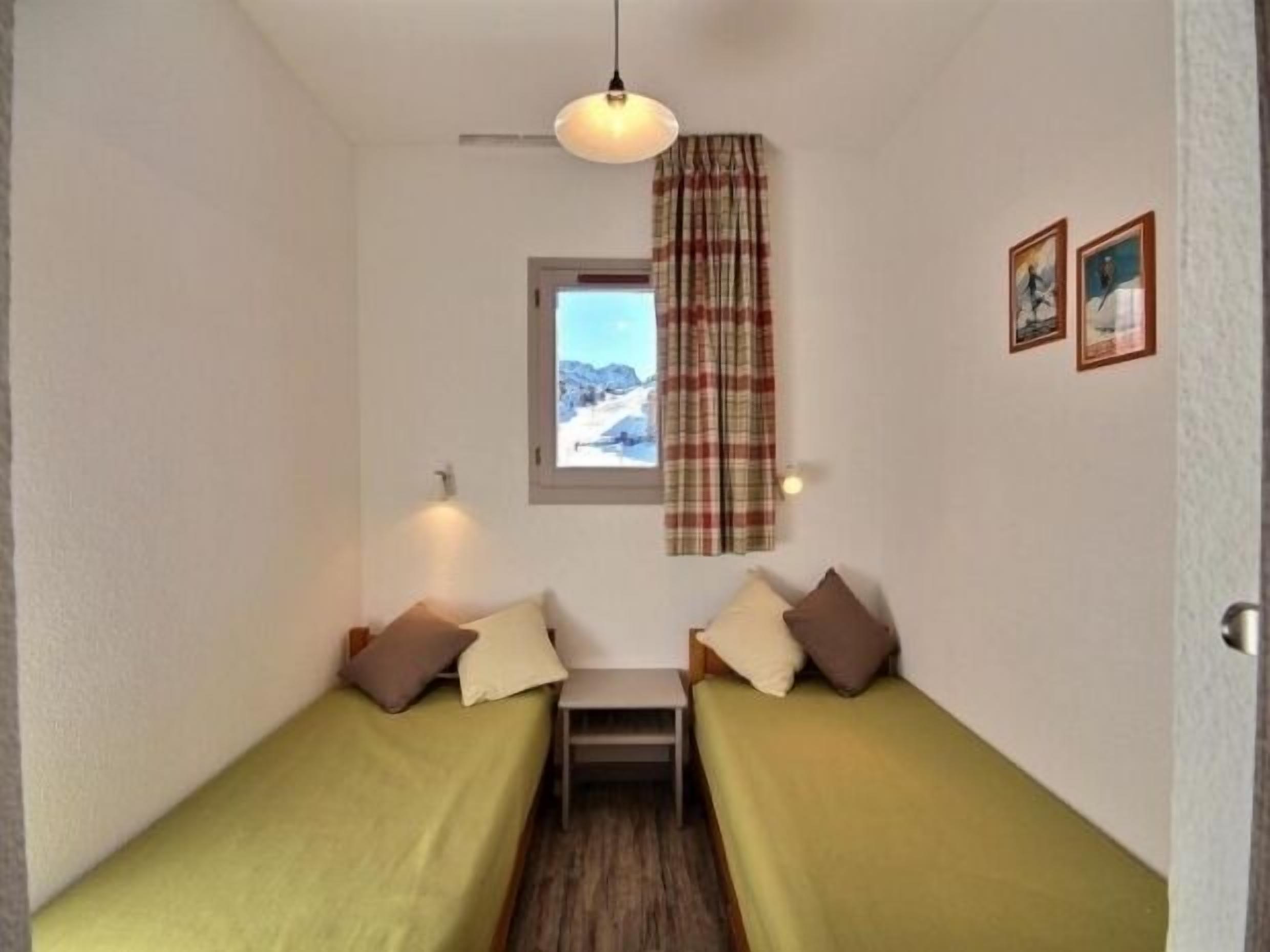 Un petit logement composé d’une seule pièce, avec deux lits simples, une fenêtre donnant sur un paysage enneigé, un luminaire suspendu et deux tableaux encadrés accrochés au mur.