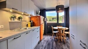 Een kleine keuken met een houten eettafel en stoelen, een inbouwoven, en een raam met uitzicht op de omgeving.