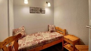 Een kleine, gezellige kamer met een eenpersoonsbed, een houten stoel en een tafeltje. Twee wandlampen en drie ingekaderde foto’s hangen boven het bed.