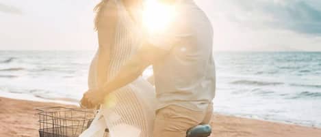 Una pareja en bicicleta con una cesta, pedaleando en una playa con el océano al fondo.