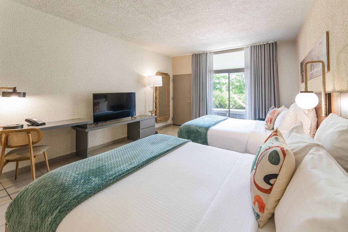 A hotel room with two beds, a desk with a TV, a chair, and a window with curtains.