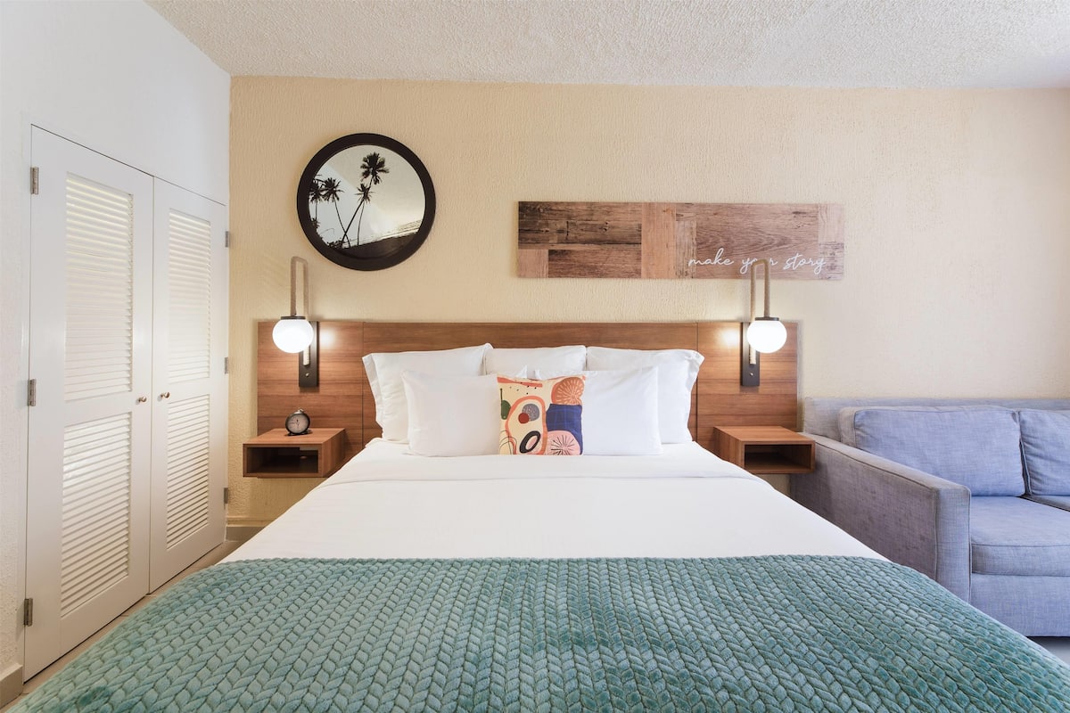 A hotel room with a bed, a nightstand, a wall clock, a mirror, and a wall art piece.