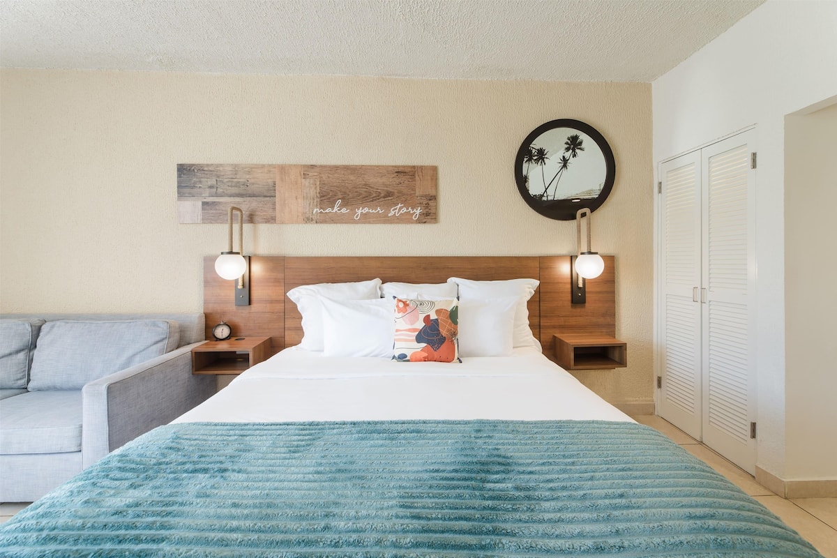 A hotel room with a large bed, a nightstand, a bedside table, a wall clock, a mirror, and a wardrobe.