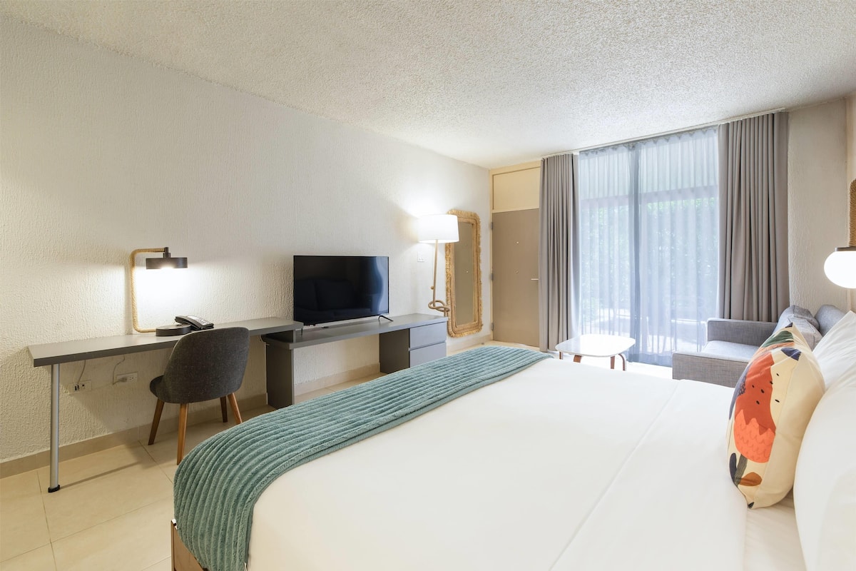 A hotel room with a bed, a desk with a TV, a chair, a small table, and a window with curtains.