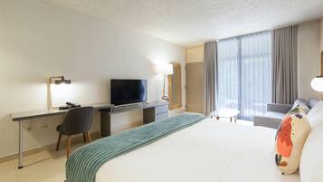 A hotel room with a bed, a desk with a TV, a chair, a small table, and a window with curtains.
