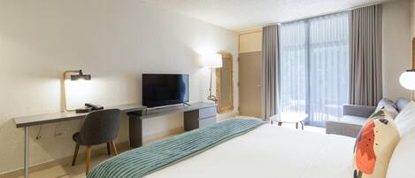 Quarto de hotel com cama, escrivaninha com televisão, cadeira, mesinha e janela com cortinas.