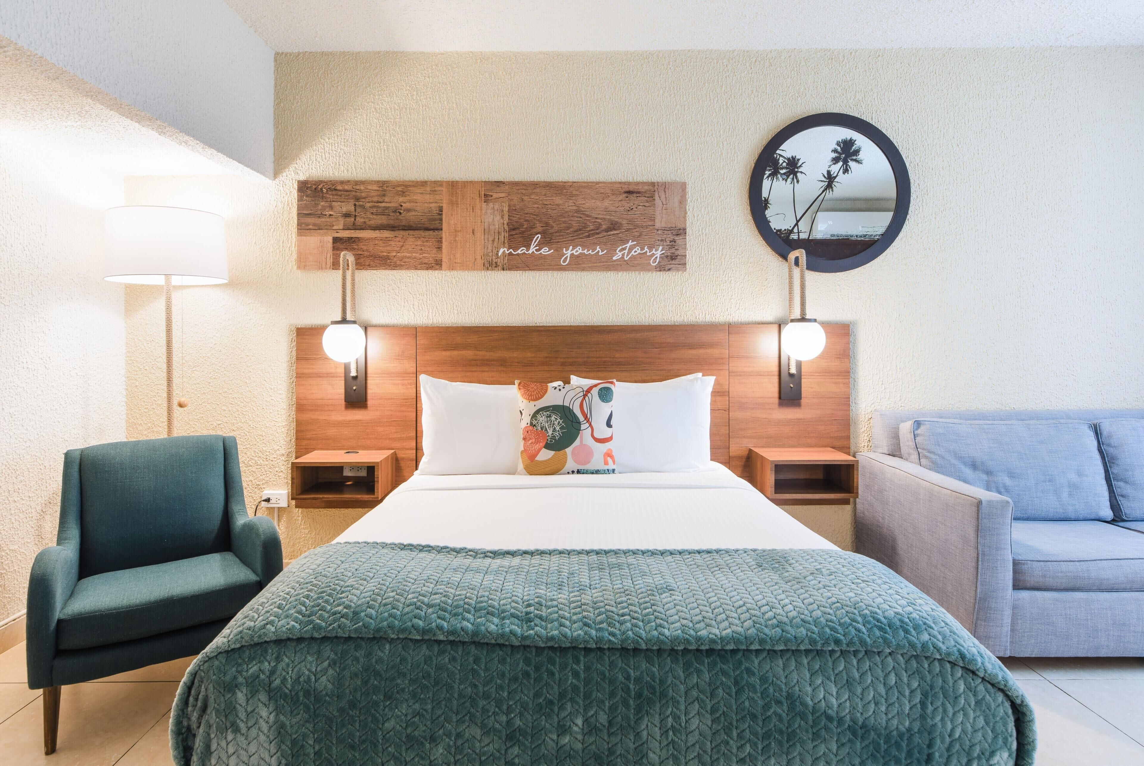 Ein Hotelzimmer mit einem Bett, einem blauen Sessel, einem grauen Sofa, einem wandmontierten Spiegel und einer dekorativen Holzverkleidung mit dem Schriftzug 'make your story'.