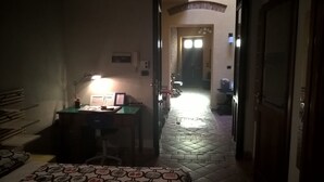 En svagt upplyst hall leder till ett rum med ett skrivbord, en stol och en lampa. Det ligger en mönstrad matta på golvet, och det finns en dörr på höger sida.
