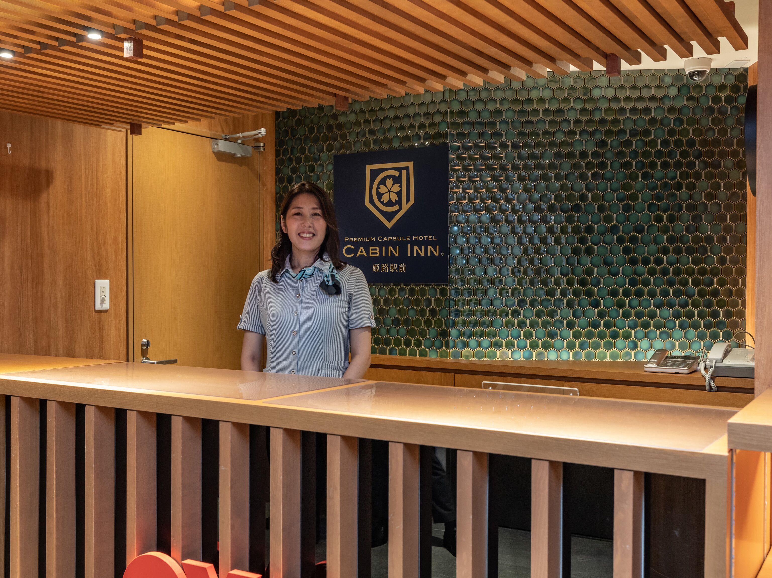 A reception desk with a wooden finish, a sign for the Premium Capsule Hotel Cabin Inn, and a wall with a honeycomb pattern.