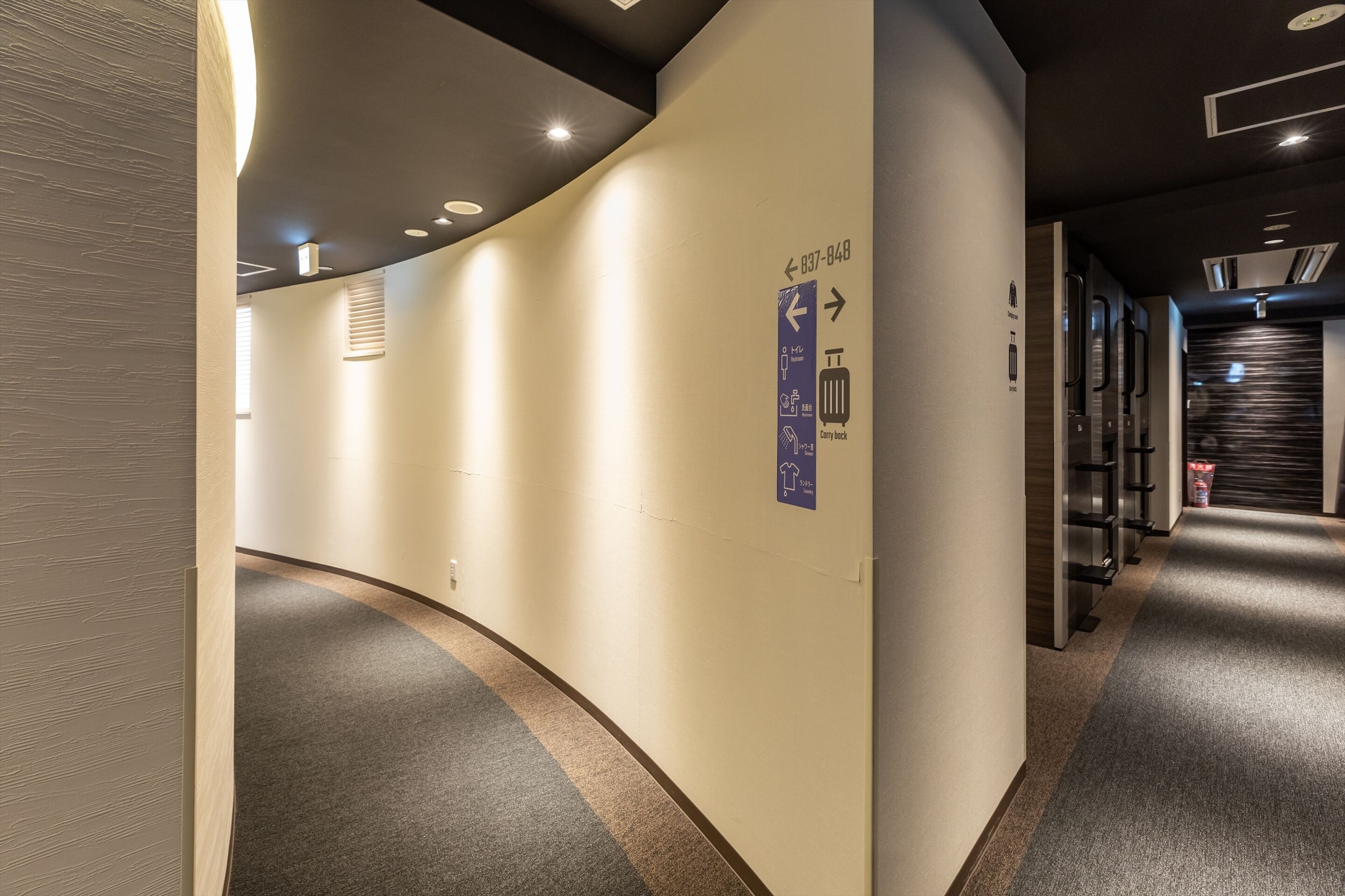 A modern hotel corridor with directional signs, a luggage storage area, and a well-lit ceiling.