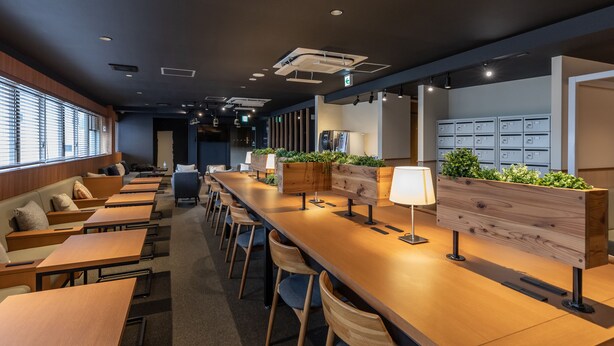 A modern, well-lit office space with long tables, chairs, and a reception area with a wooden counter and plants. - Tabist CapsuleHotel APODS Himeji Station (Himeji)