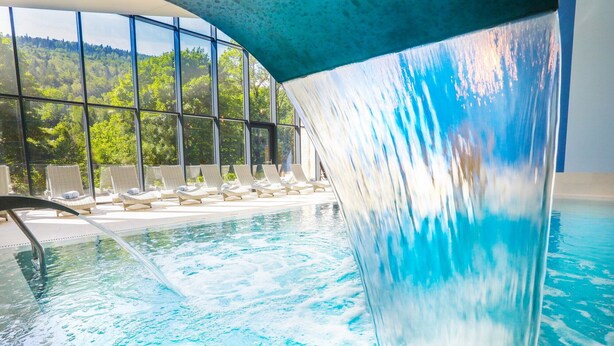 Una piscina con cascata, sedie a sdraio e vista su una foresta attraverso ampie finestre.
