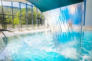 Una piscina con cascada, sillas de descanso y vistas a un bosque a través de amplios ventanales.