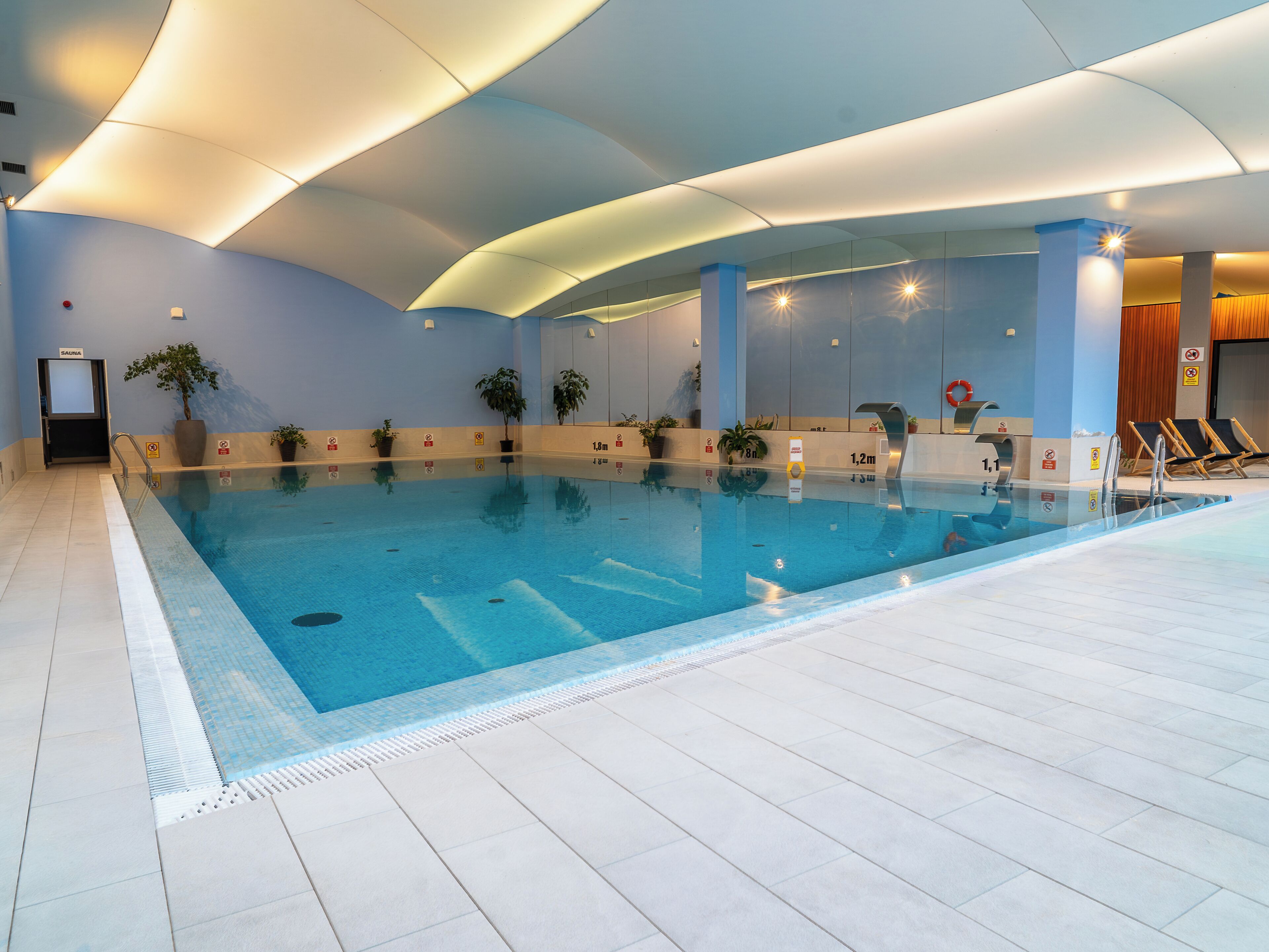 Une piscine intérieure à l’eau claire, entourée d’un sol carrelé, de plantes en pot et d’un plafond doté d’un éclairage encastré.