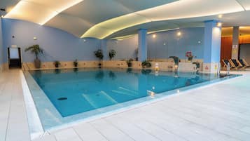 An indoor swimming pool with clear water, surrounded by tiled flooring, potted plants, and a ceiling with recessed lighting.