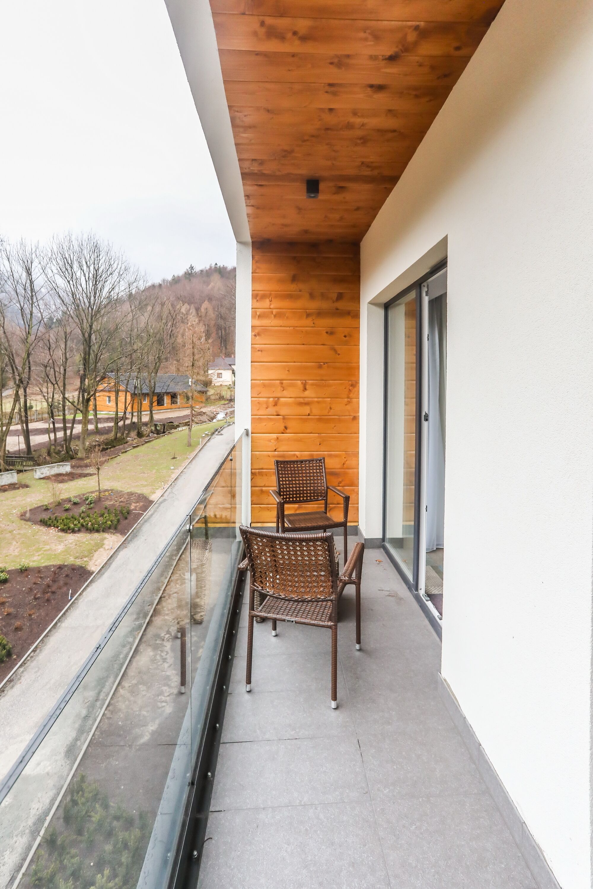 Un balcon doté d’un plafond en bois, d’une chaise et d’une balustrade en verre, offrant une vue sur un paysage arboré et un bâtiment.