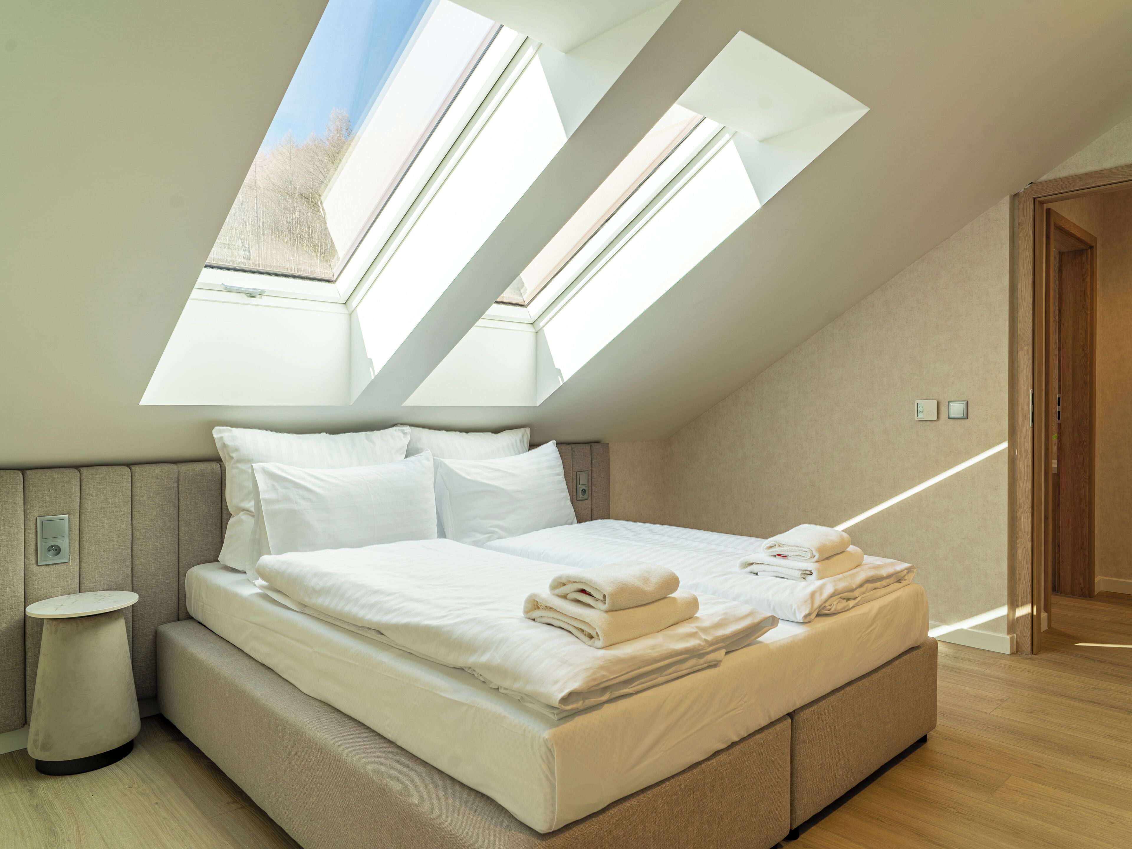 Un dormitorio con tragaluz, una cama con ropa blanca, una mesita de noche y suelo de madera.