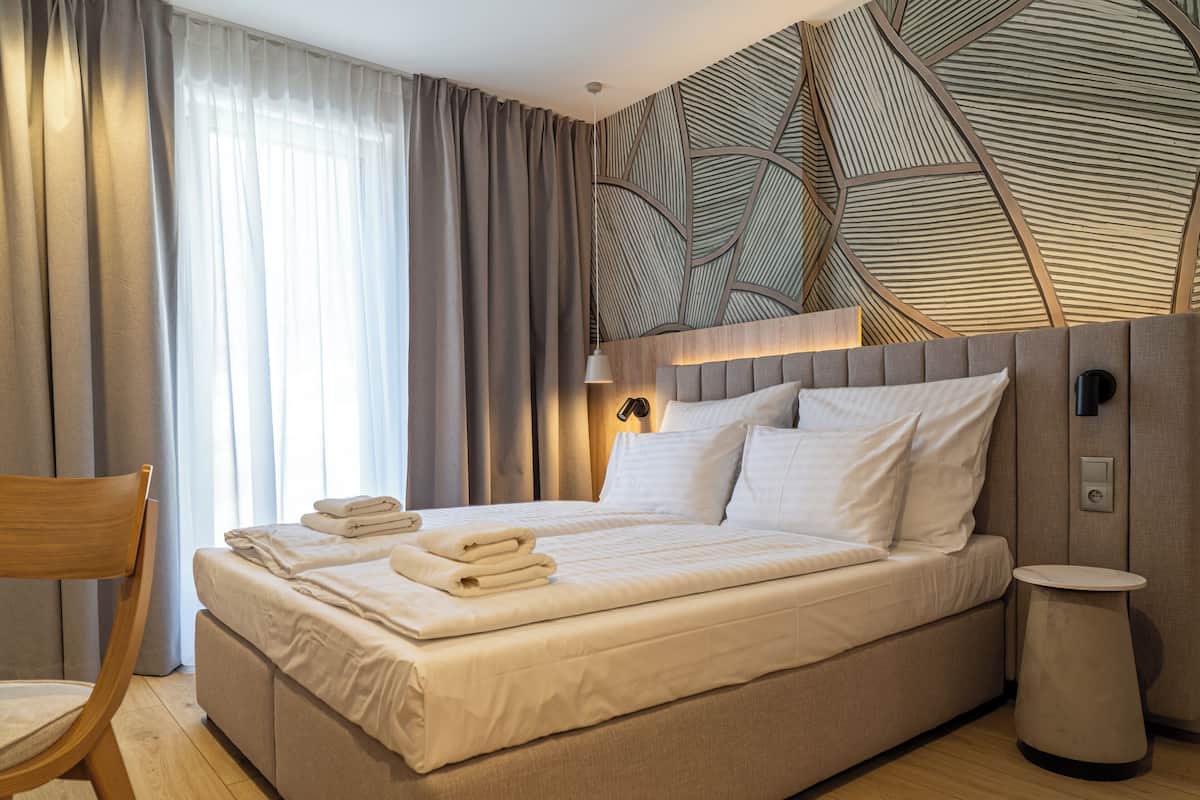 A modern hotel room with a large bed, a wooden chair, a bedside table, and a decorative wall panel.