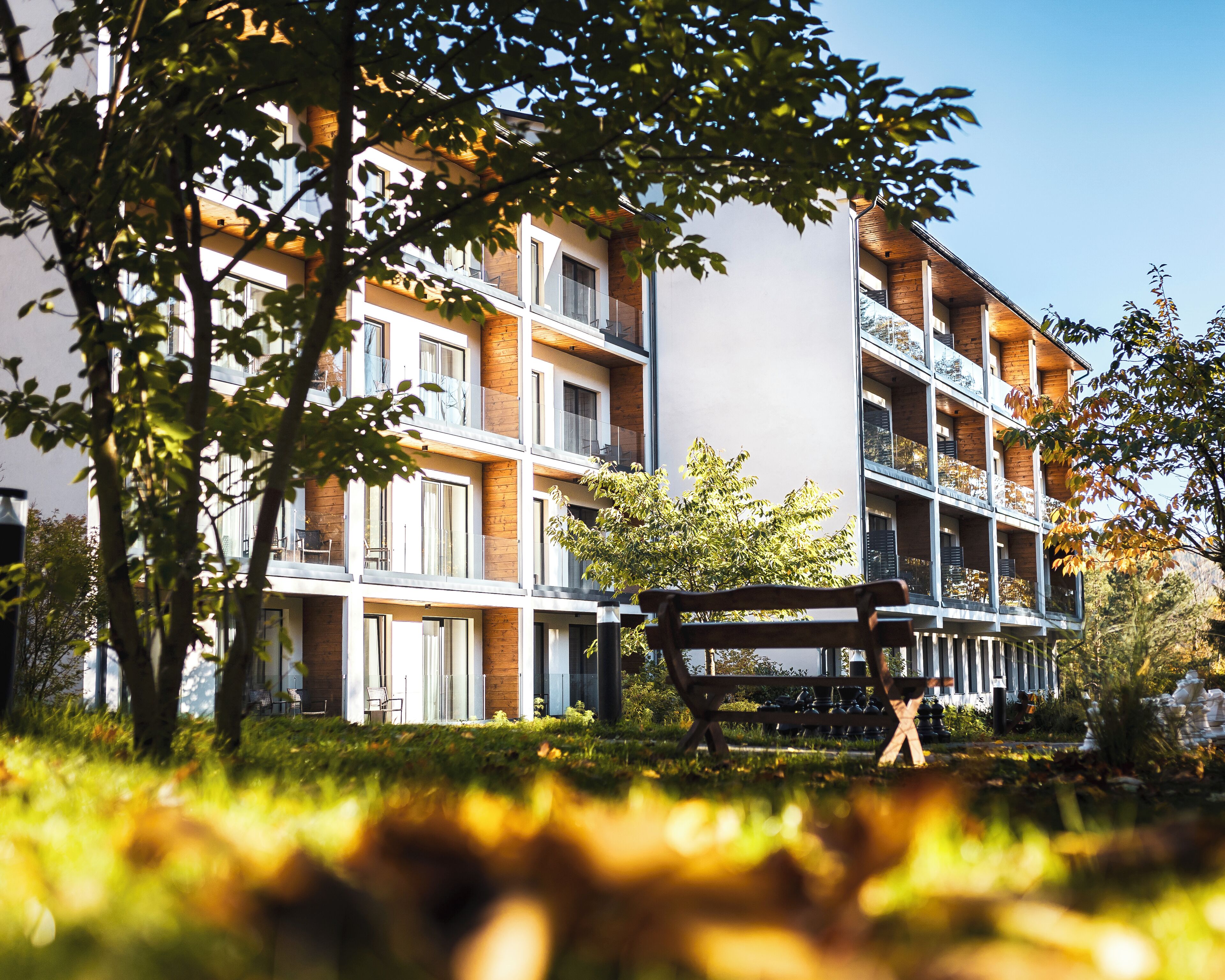 A multi-story residential building with balconies, a grassy area in the foreground, and a bench.