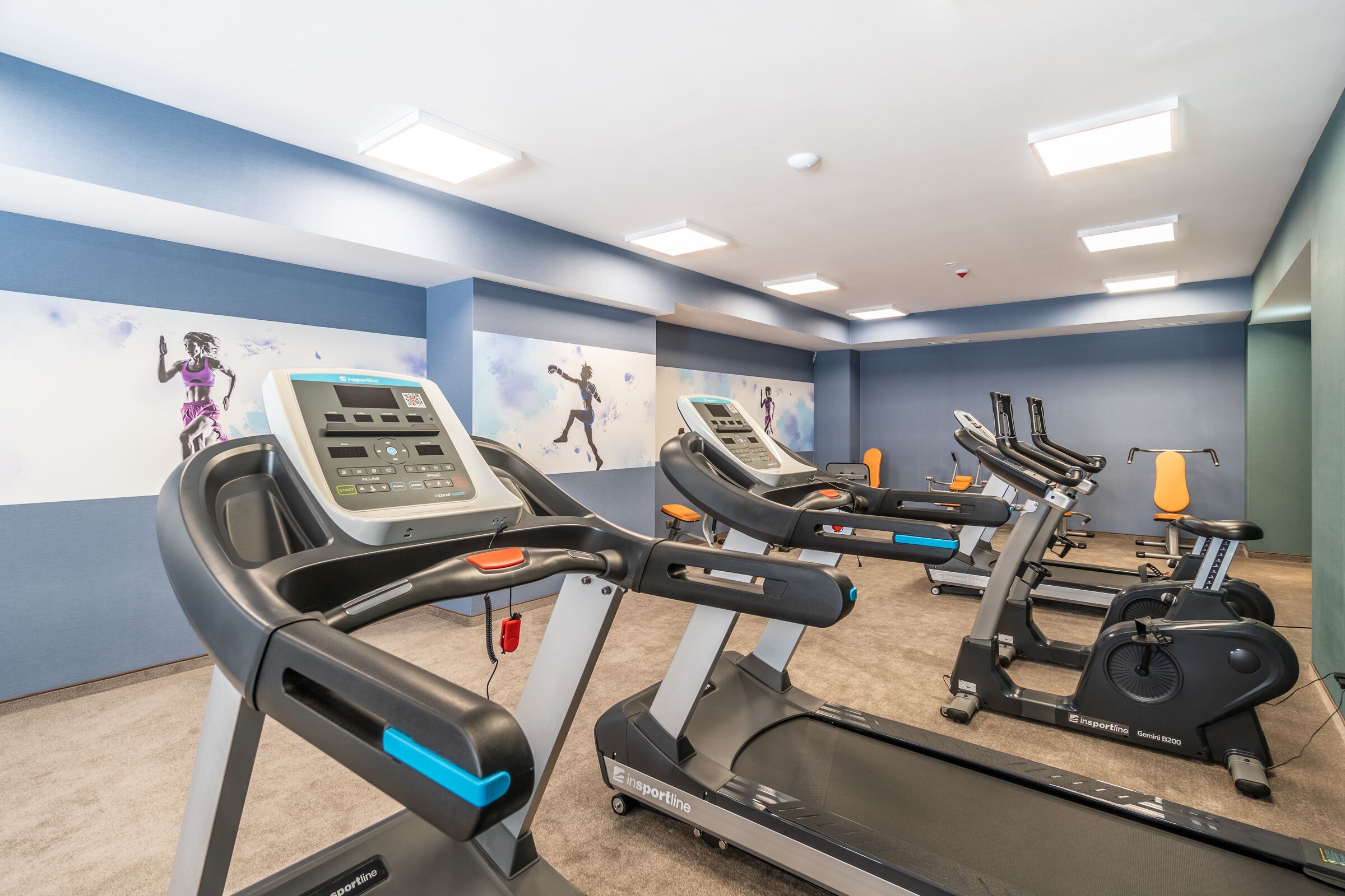Un gimnasio con varias cintas de correr y bicicletas estáticas, equipado con pantallas digitales y monitores de frecuencia cardíaca.