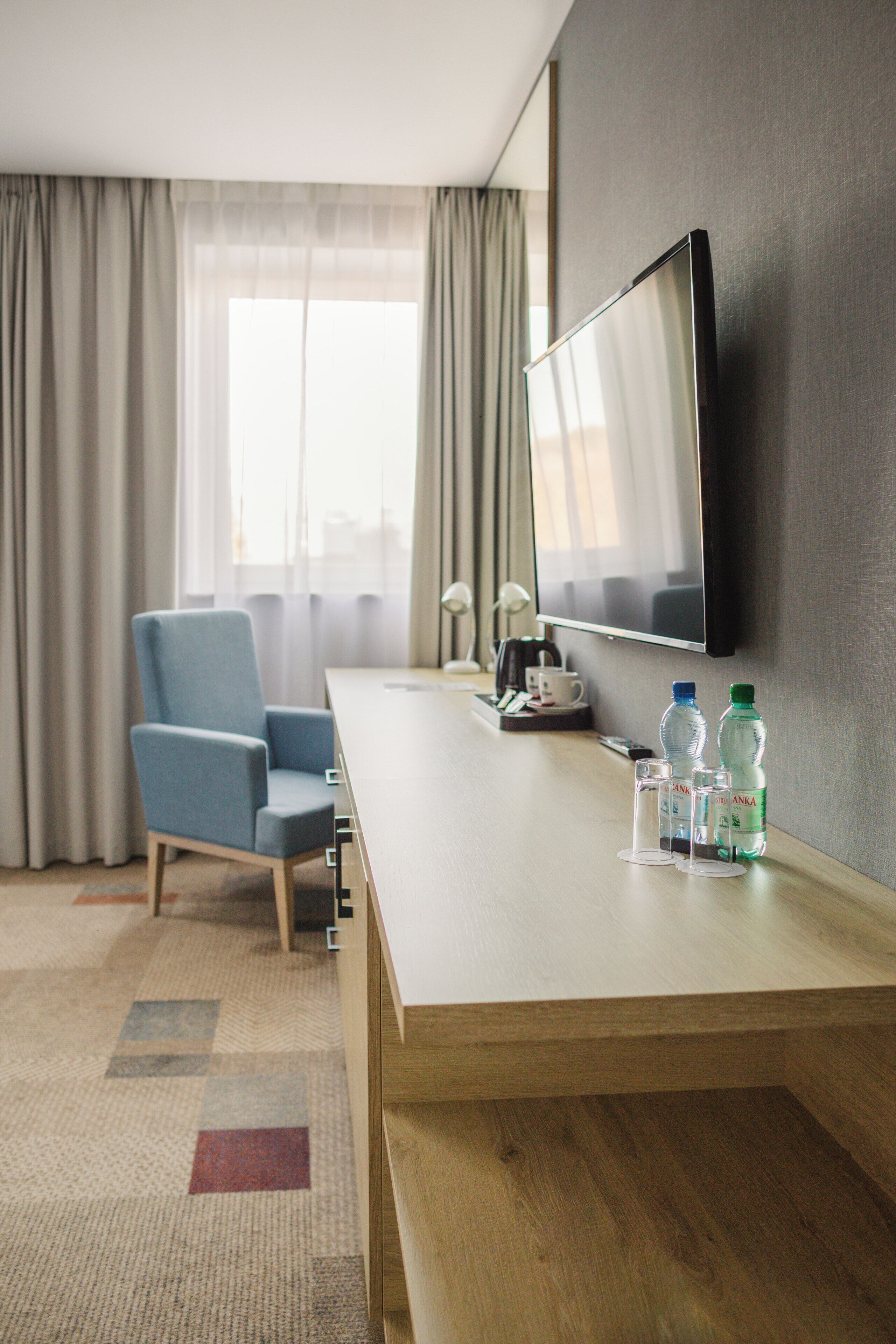 A hotel room with a wooden desk, a flat-screen TV, a blue chair, a coffee maker, and two water bottles.