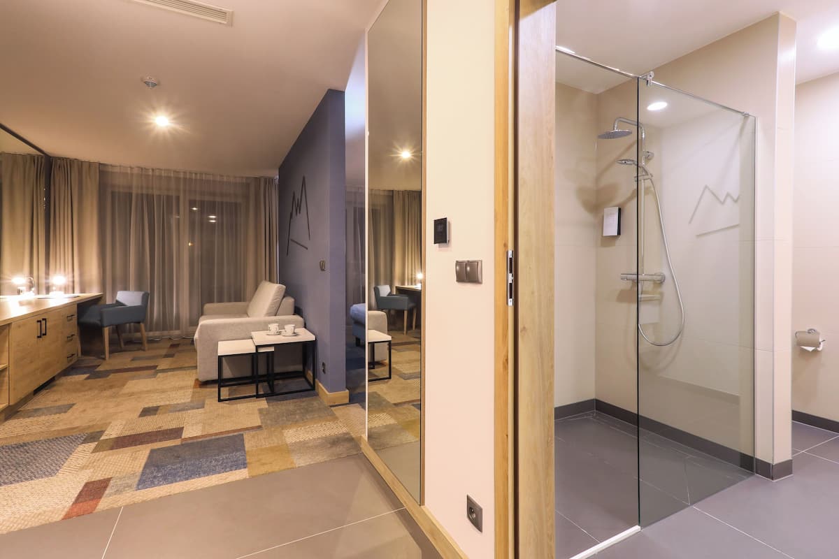 A modern hotel room with a glass-enclosed shower, a sofa, a small table, and a view of the dining area through a glass partition.