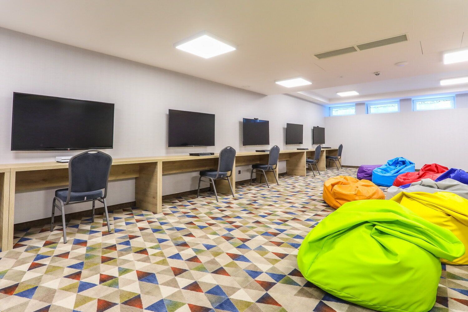 Una habitación con varias estaciones de trabajo, cada una con una silla y un monitor de pantalla plana. Hay pufs de distintos colores en el suelo.