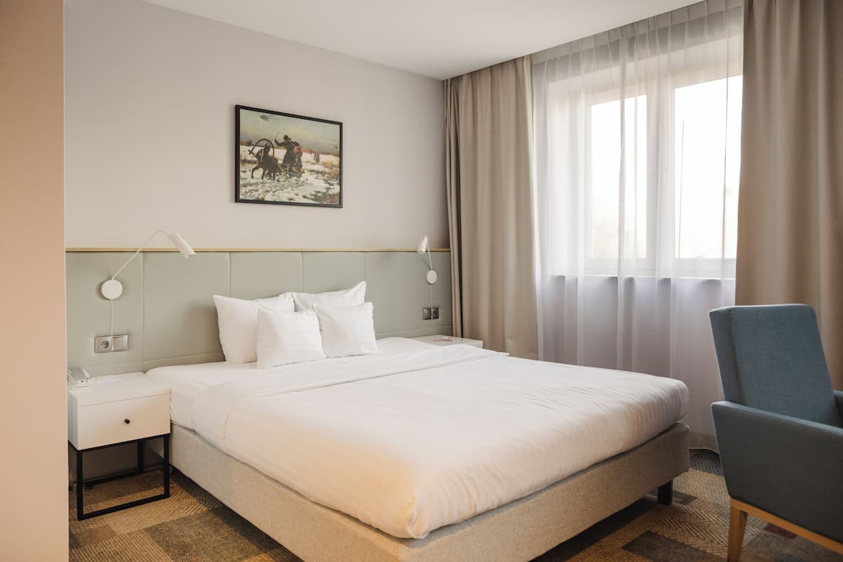 A hotel room with a large bed, a bedside table, a chair, a painting on the wall, and a window with curtains.