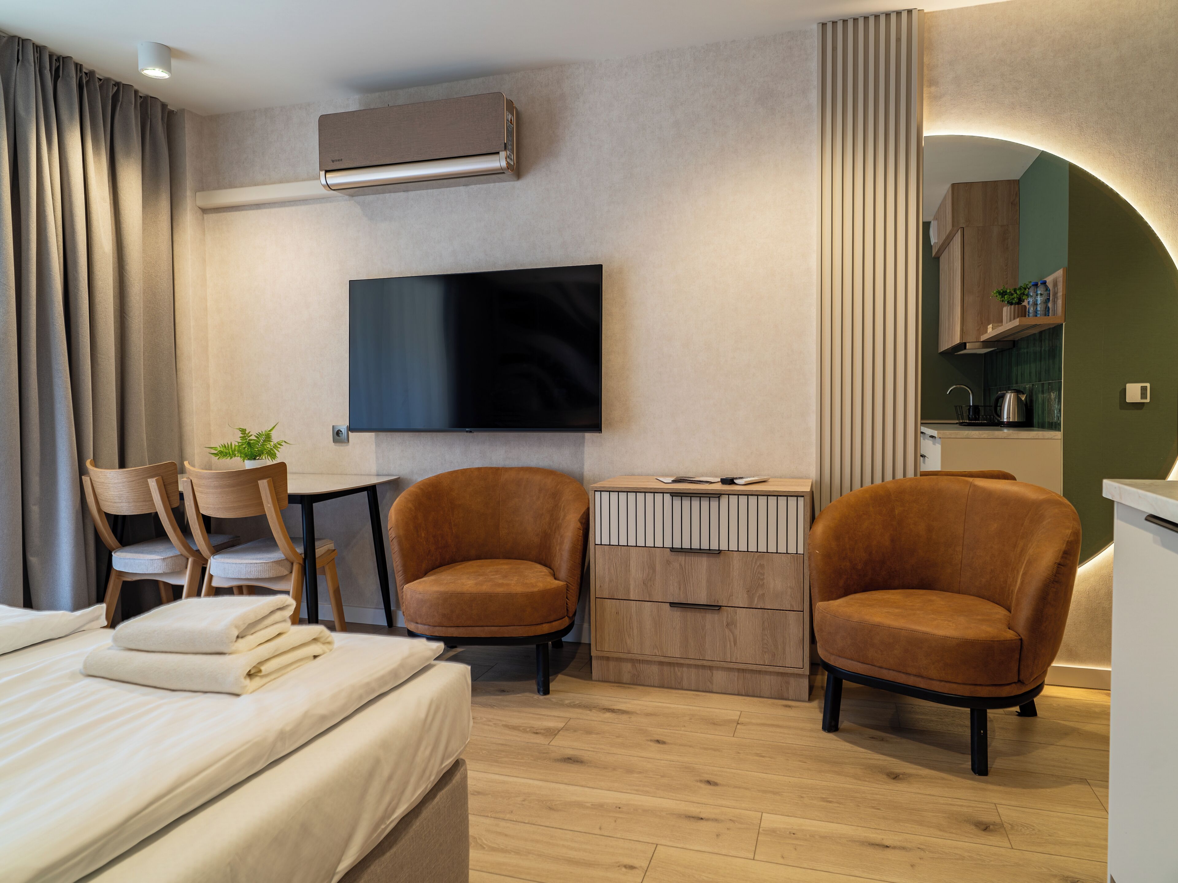 A modern hotel room with a bed, two armchairs, a TV, a small table, and a kitchenette.