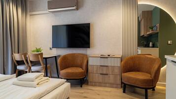 A modern hotel room with a bed, two armchairs, a TV, a small table, and a kitchenette.