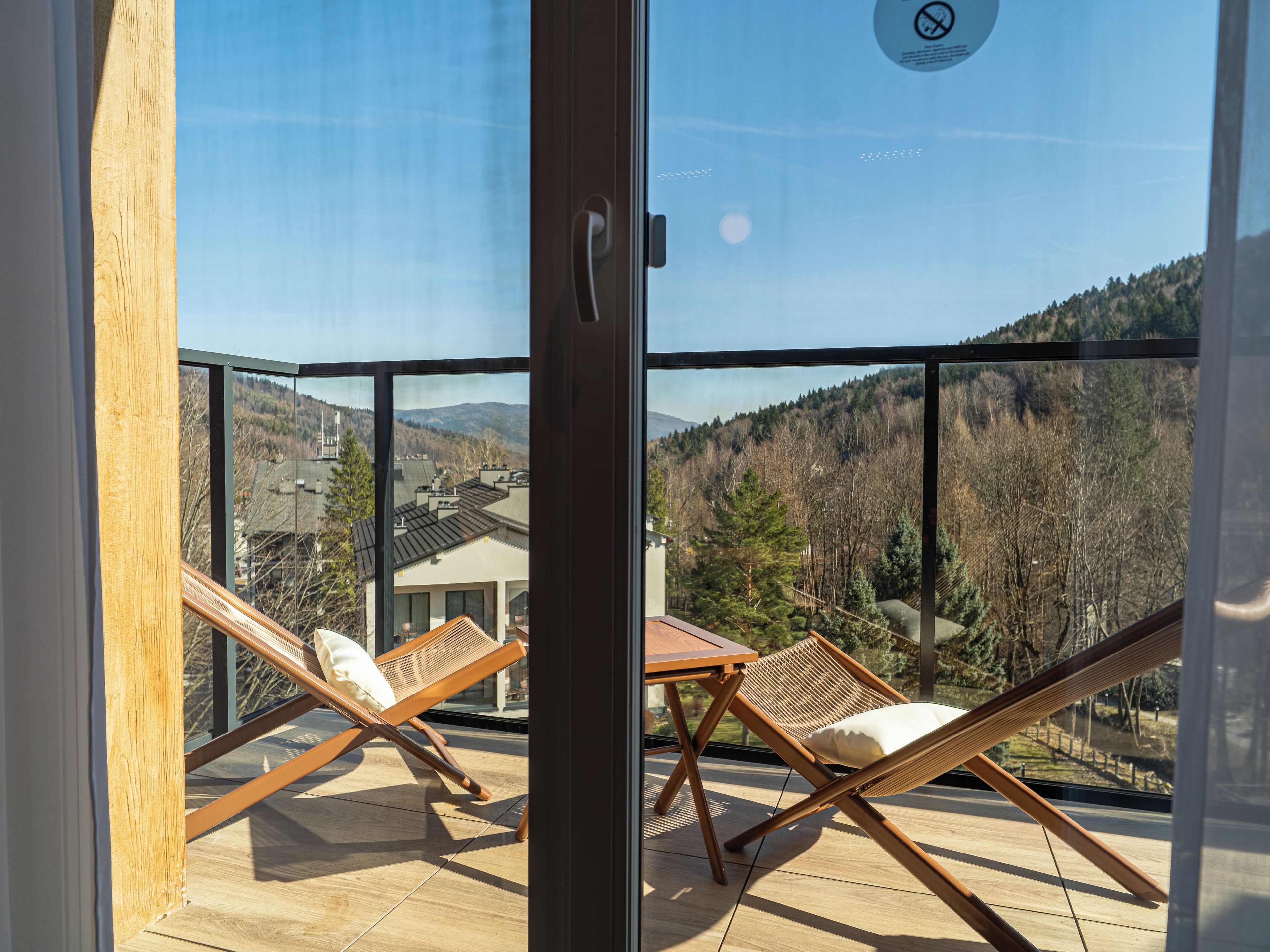 Een balkon met twee houten loungesteunen en een tafel, met uitzicht op een bebost glooiend landschap en een woongebied.