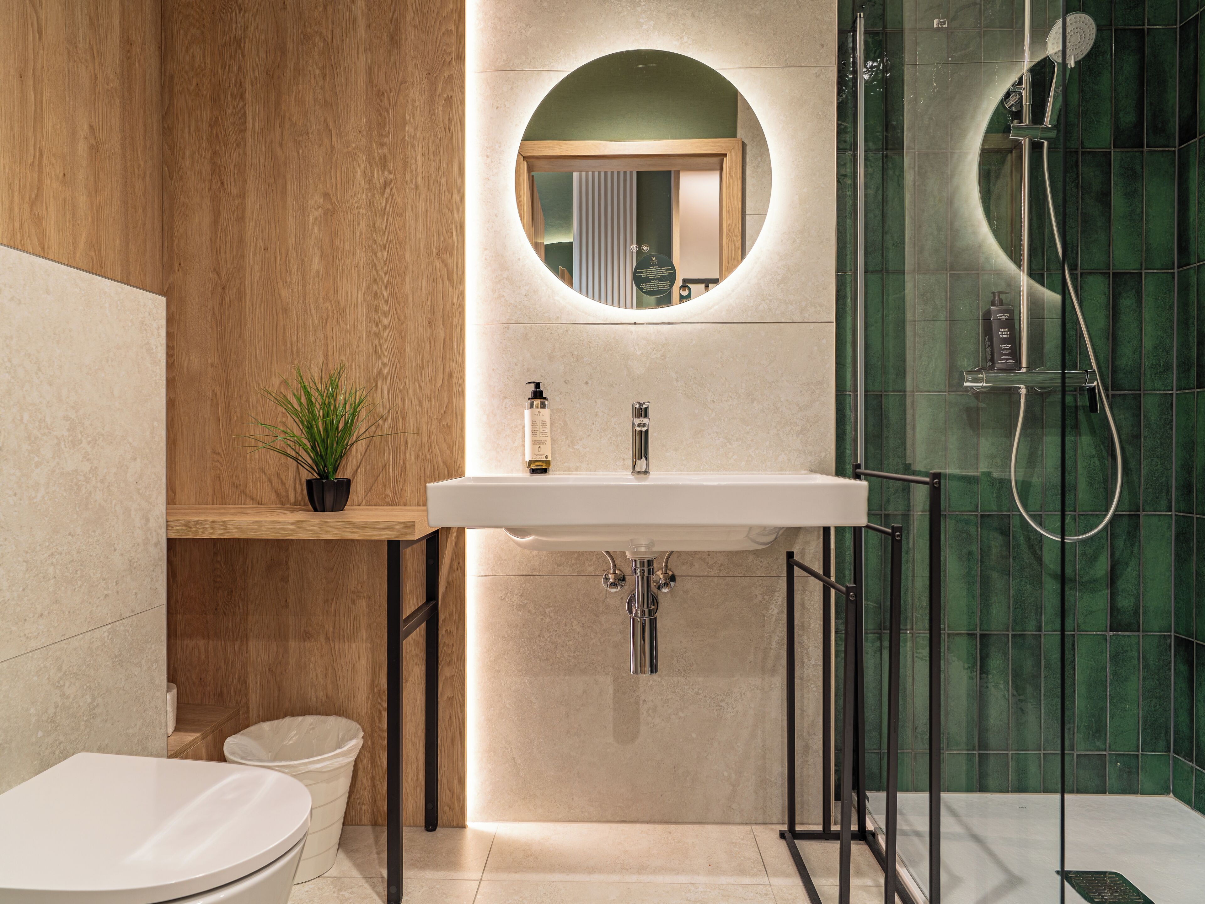 A modern bathroom with a wooden vanity, a round mirror, a white sink, a toilet, and a shower area.
