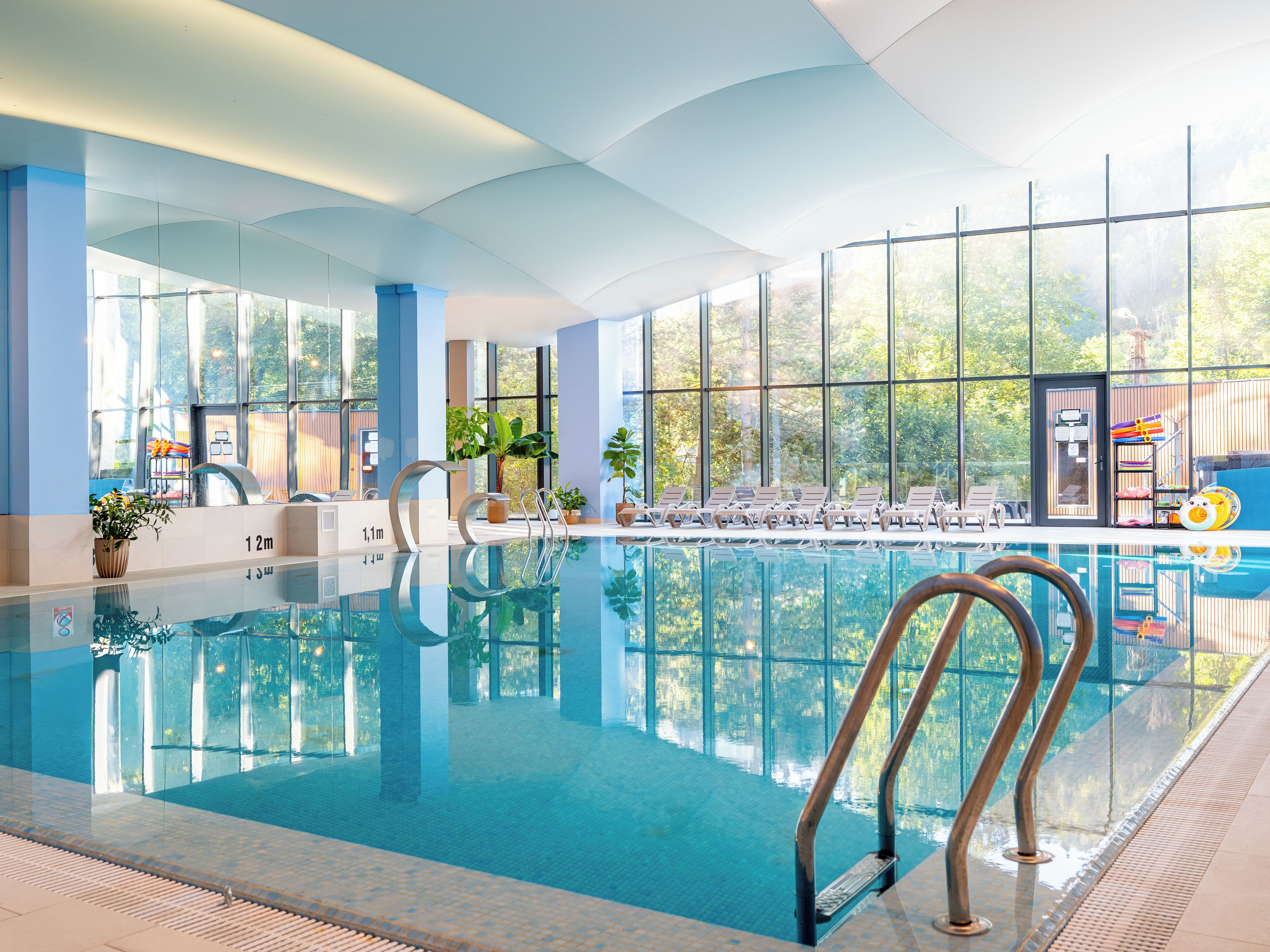 Une piscine intérieure avec des parois en verre transparent, des chaises longues et une vue sur l’extérieur.