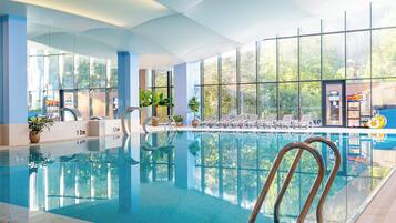 Une piscine intérieure avec des parois en verre transparent, des chaises longues et une vue sur l’extérieur.