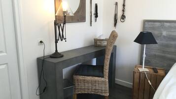 Una habitación pequeña con escritorio, una silla de mimbre, cama y mesita de noche con lámpara.