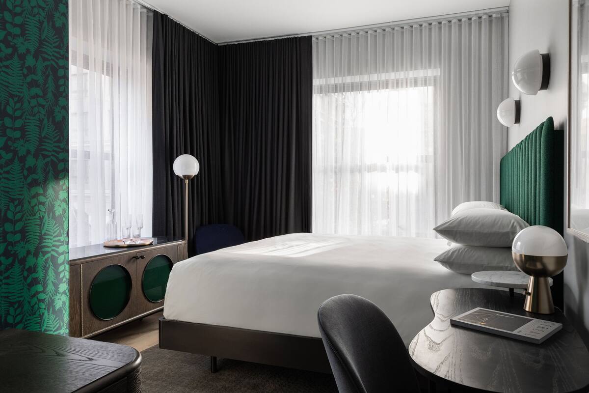 A modern hotel room with a large bed, a bedside table with a lamp, a chair, and a desk with a computer.