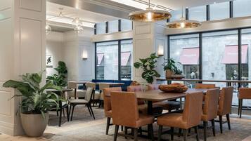 A spacious dining area with a large table and chairs, potted plants, and large windows with view of the city.