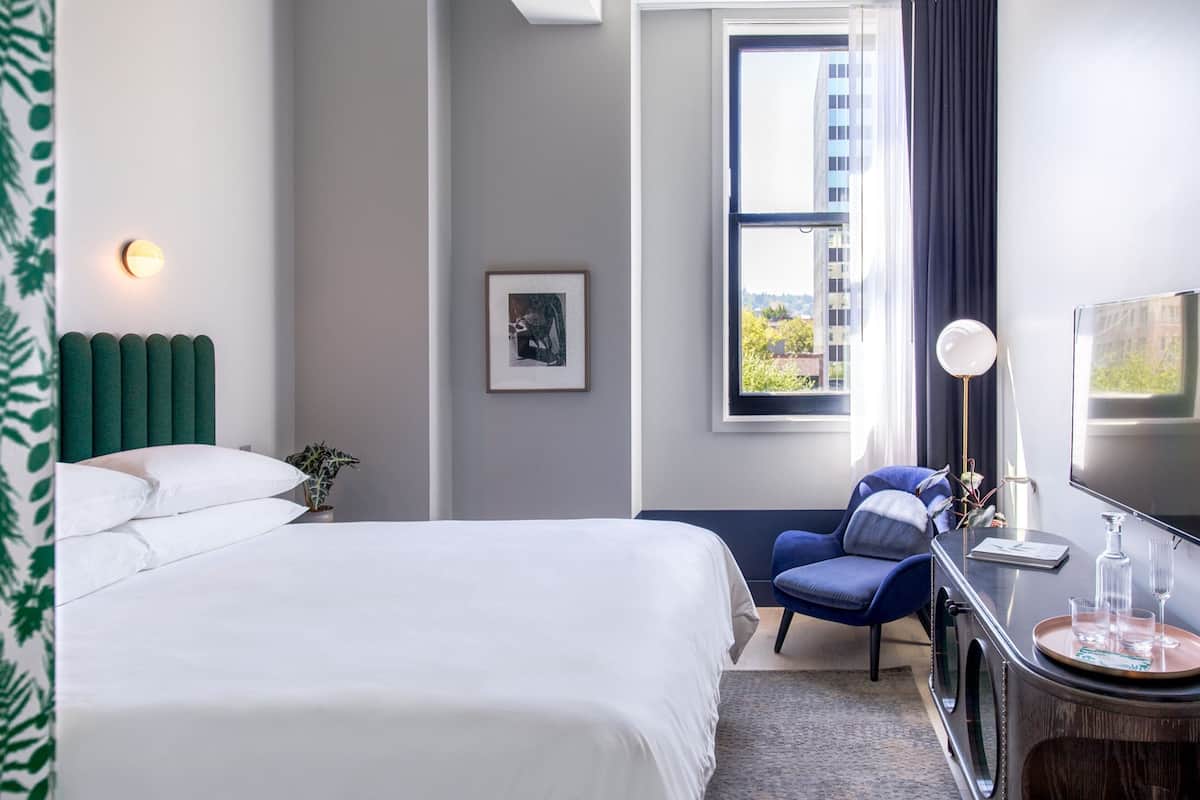 A modern hotel room with a large bed, a desk with a flat-screen TV, a chair, and a window with a view of buildings and trees.