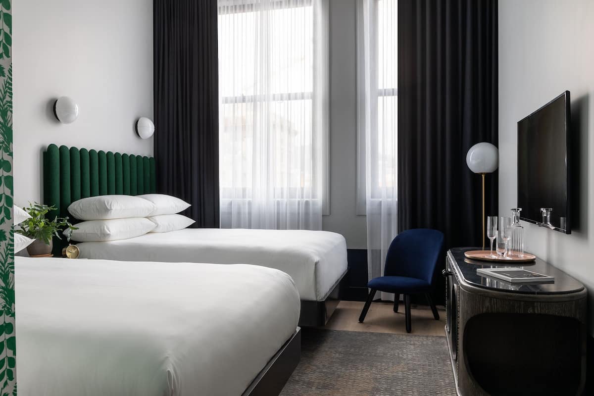 A hotel room with two beds, a desk with a chair, a TV, and a window with curtains.
