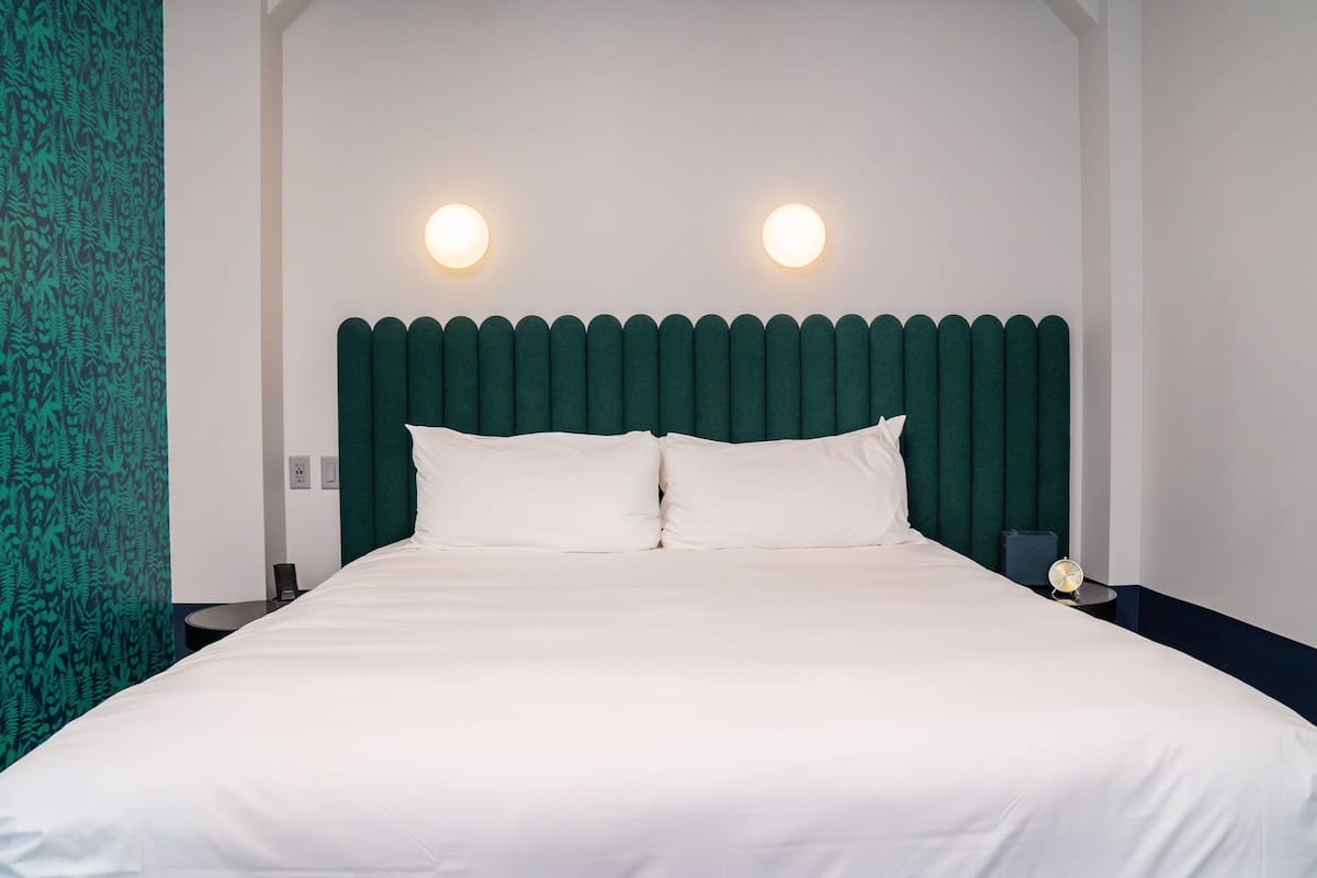 A neatly made bed with white linens, a green upholstered headboard, and two wall-mounted lights.