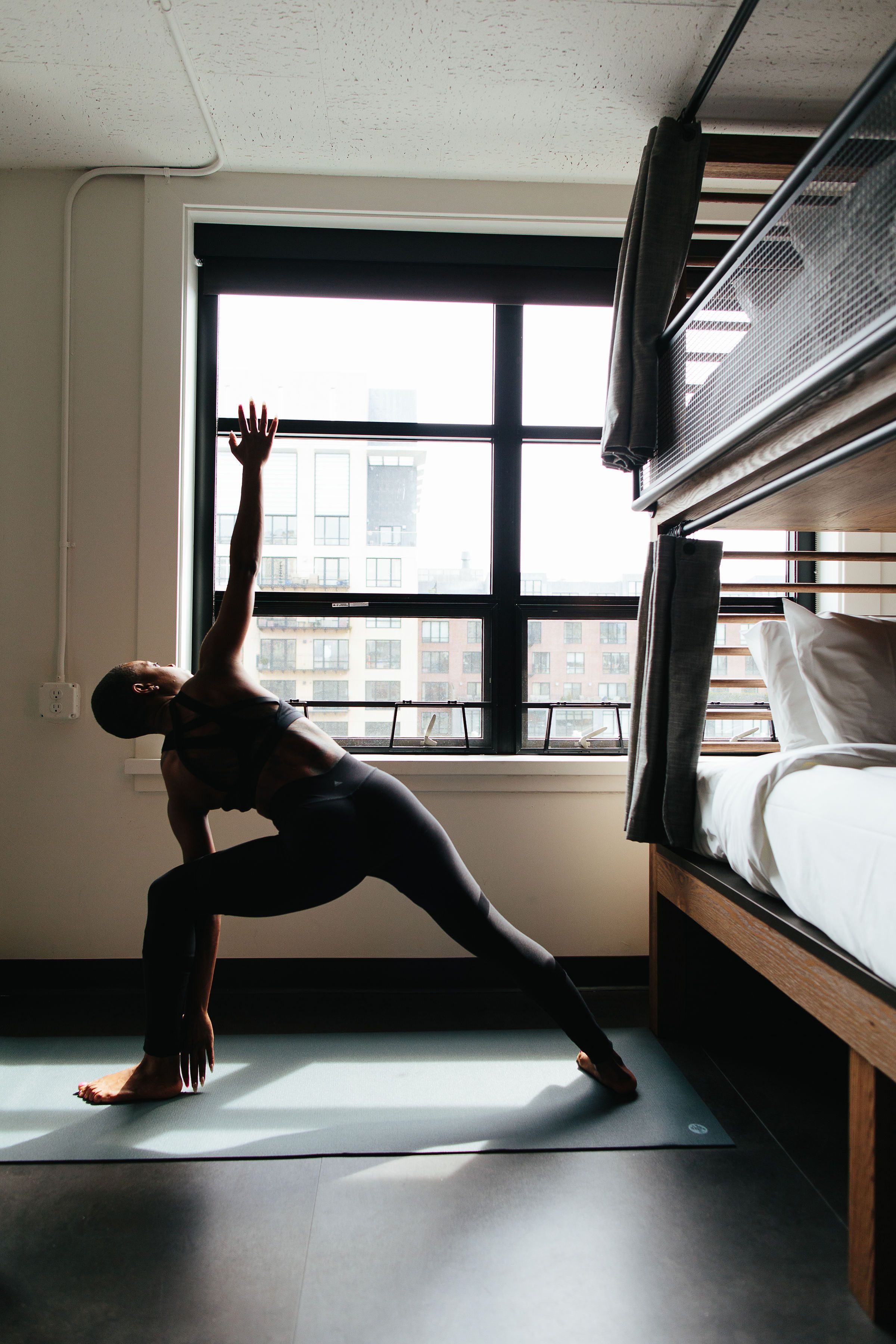 A person performing a yoga pose on a mat in a room with a bunk bed and a window with a view of buildings.