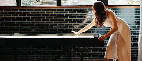 Una mujer apoyada en una mesa en una habitación con paredes de ladrillo y una ventana.