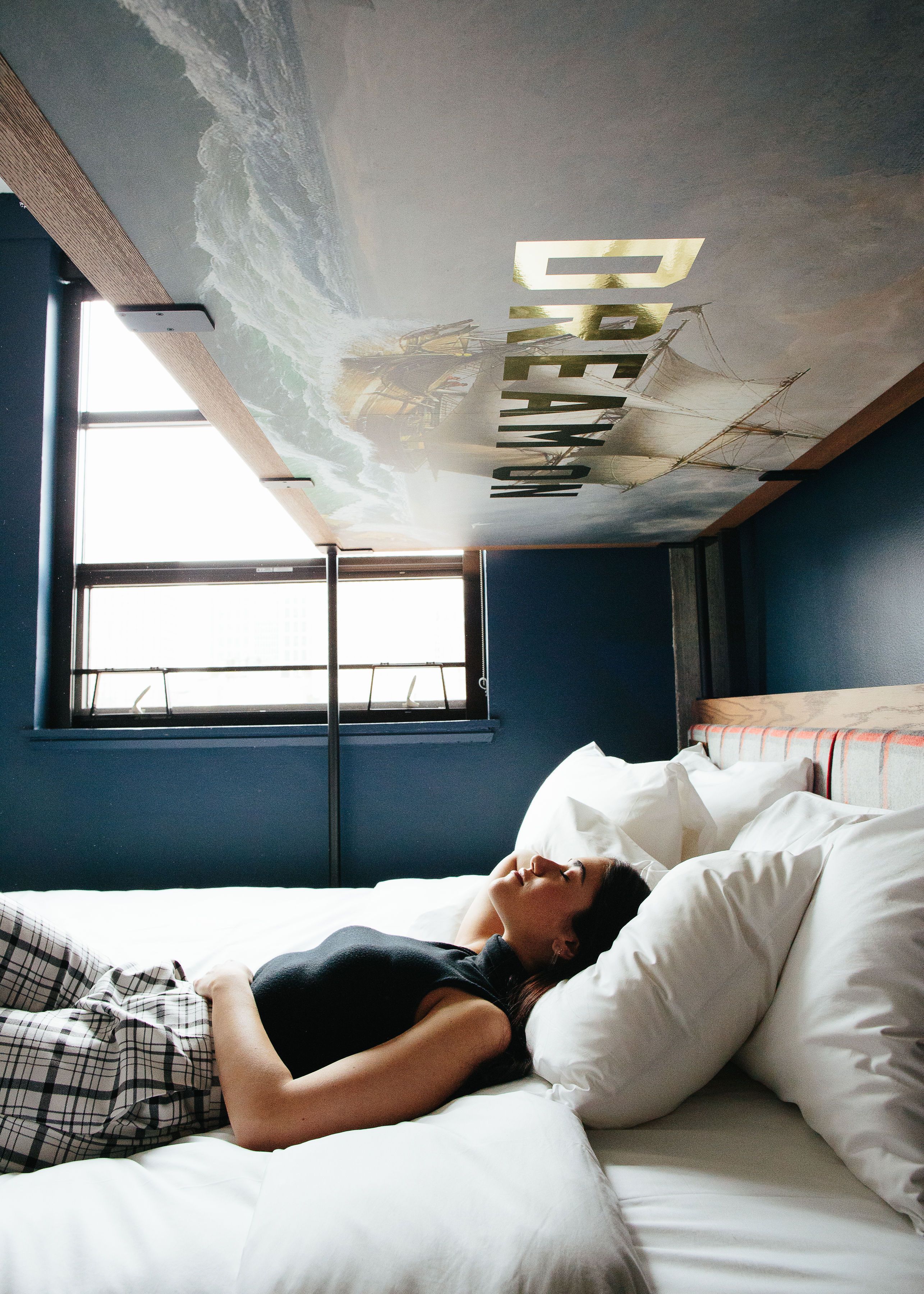 Une personne allongée sur un lit dans une pièce dotée d’une grande fenêtre, d’une peinture murale au plafond et d’une tête de lit en bois.
